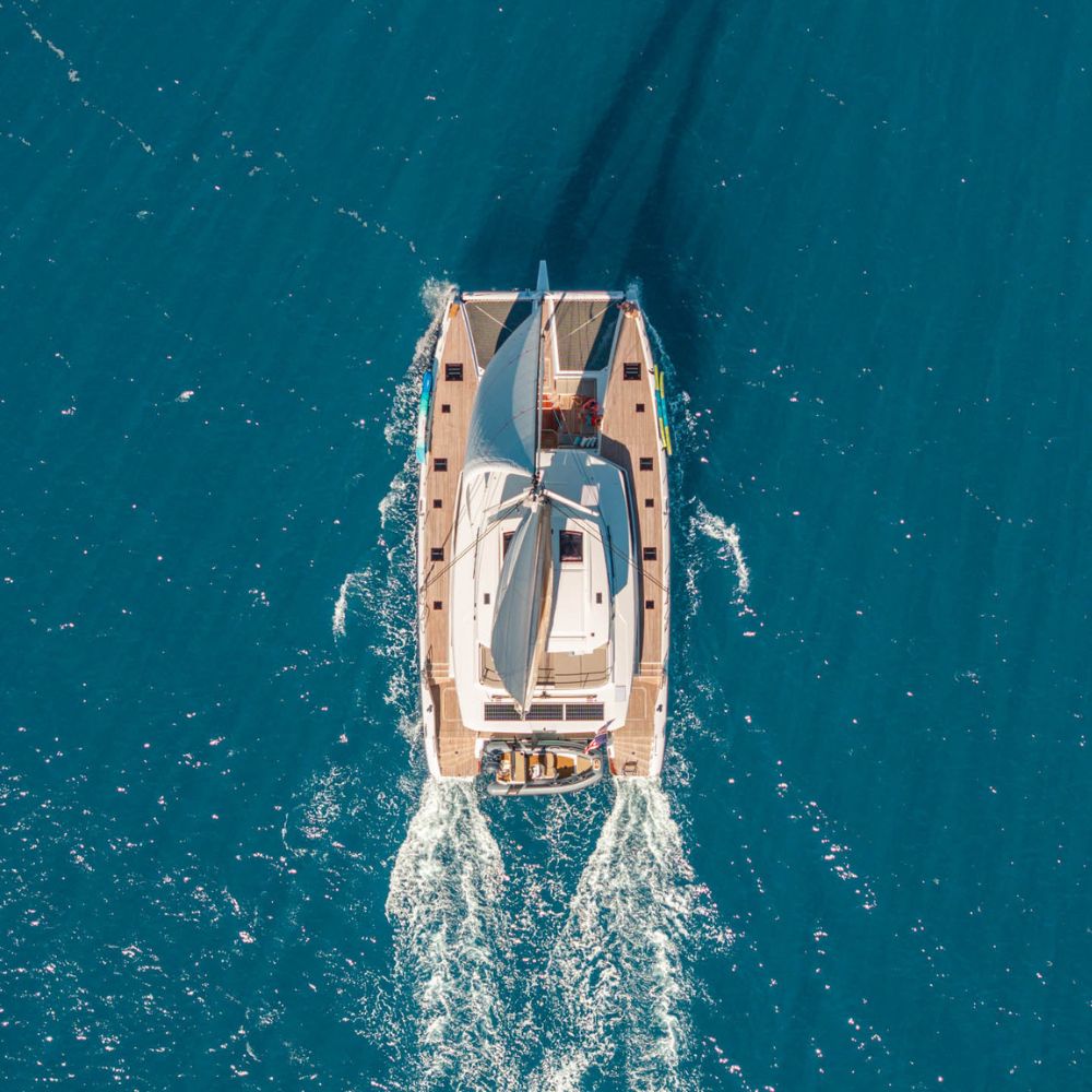 Overhead drone view of a sailing catamaran underway on vivid blue water, with teak decks, sails up and a tender in tow